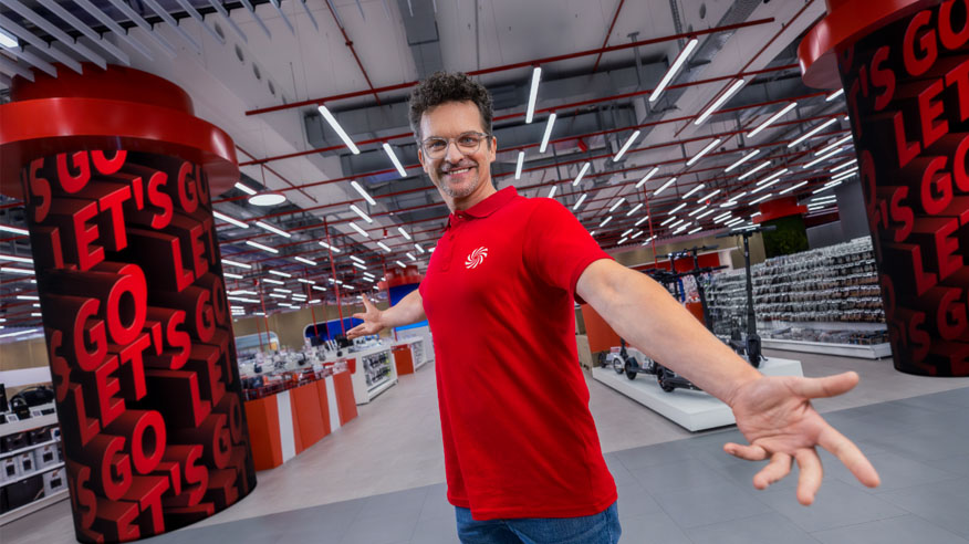 Mężczyzna w czerwonej koszulce stoi w sklepie z kolumnami z napisem "Let's Go". W tle półki z towarem i oświetlenie.