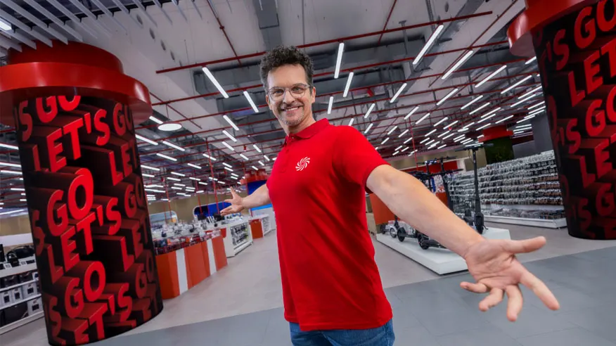 Mężczyzna w czerwonej koszulce stoi w sklepie z kolumnami z napisem "Let's Go". W tle półki z towarem i oświetlenie.