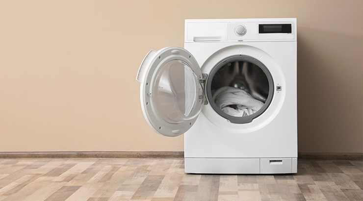 Pralka automatyczna, biała, z otwartymi drzwiami, stoi na podłodze imitującej drewno, na tle beżowej ściany. W środku widać pranie.