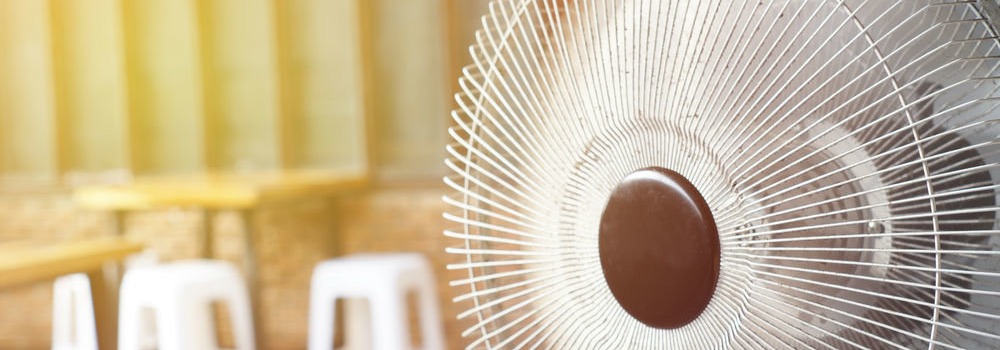 Na pierwszym planie biały wentylator z brązowym środkiem, w tle stolik i białe krzesła.