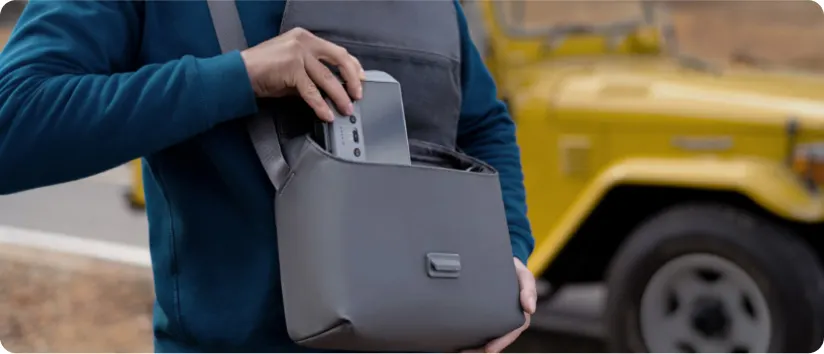 Osoba wkłada szary przedmiot do szarej torby na ramię. W tle widać żółty samochód terenowy.