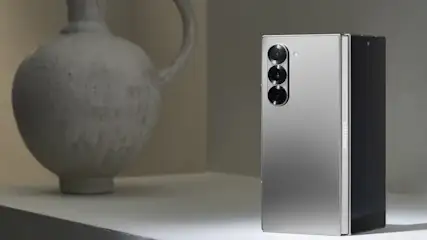 Szary, składany smartfon z trzema obiektywami aparatu obok szarego wazonu na blacie.