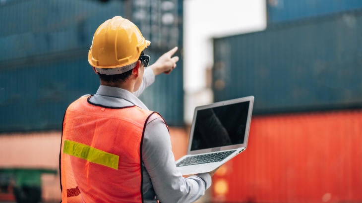 Osoba w żółtym kasku i pomarańczowej kamizelce trzyma laptopa i wskazuje na kontenery w tle.