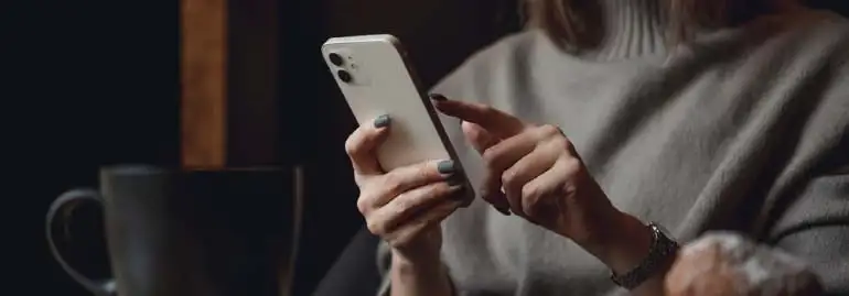 Kobieta w swetrze obsługuje smartfon. Obok stoi duży, czarny kubek. Tło jest ciemne i częściowo rozmyte.