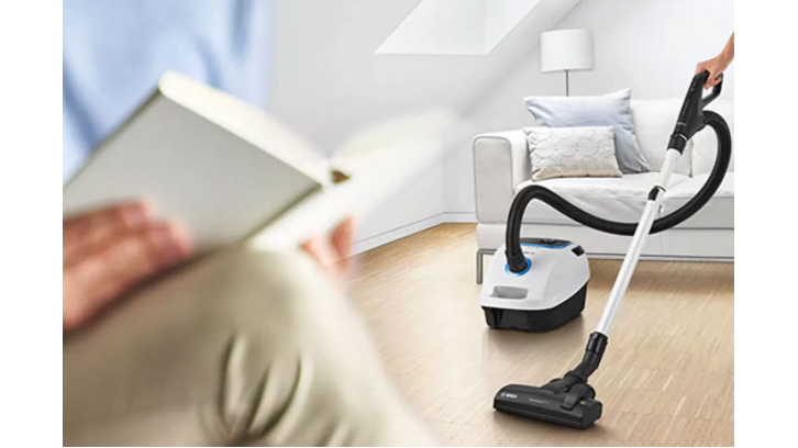 Pokój z białą sofą i lampą. Na drewnianej podłodze stoi biało-czarny odkurzacz, ktoś odkurza. Zamazana postać czyta książkę.