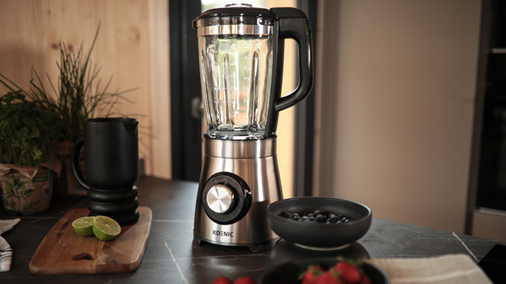 Na stole stoi srebrny blender, obok czarny dzbanek, miska z jagodami i deska z limonkami.