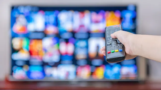 Na pierwszym planie dłoń trzyma pilota do telewizora. W tle widoczny ekran z różnymi aplikacjami w kolorach: niebieskim, pomarańczowym i białym.