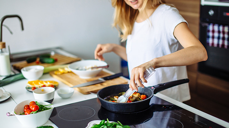 Kobieta w kuchni gotuje na płycie, używając szczypiec. Na blacie warzywa, deski do krojenia i miski. W tle piekarnik.