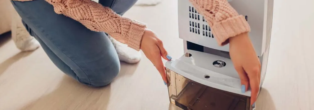 Osoba w swetrze wyjmuje pojemnik na wodę z białego osuszacza powietrza na podłodze.