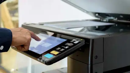 Osoba obsługuje czarne urządzenie wielofunkcyjne z dotykowym ekranem. W tle widać otwartą pokrywę i białe dokumenty wewnątrz.