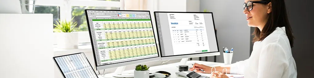Kobieta pracuje przy biurku z dwoma monitorami, laptopem, kalkulatorem i długopisem. Na monitorach arkusze kalkulacyjne i faktury.