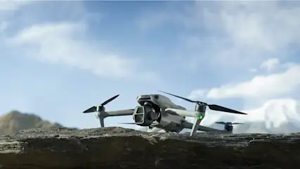 Szary dron stoi na skale. W tle widać góry i błękitne niebo z chmurami.