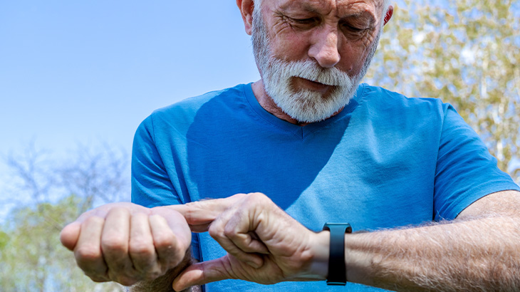 Mężczyzna w niebieskiej koszulce patrzy na czarny zegarek na nadgarstku. W tle widoczne drzewa i błękitne niebo.