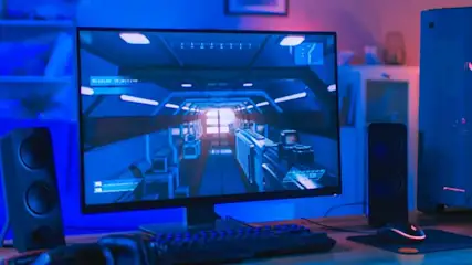 Na biurku stoi monitor z grą, klawiatura i mysz. Obok głośniki, obudowa komputera z czerwonymi wentylatorami i smartfon. W tle sofa.