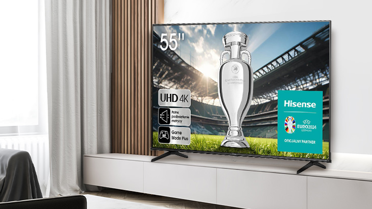 Telewizor Hisense na białej szafce prezentuje trofeum sportowe na tle stadionu. Obok logo Euro 2024 i napisy. Zasłony i drewno w tle.
