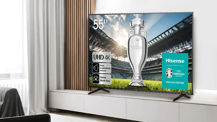 Telewizor Hisense na białej szafce prezentuje trofeum sportowe na tle stadionu. Obok logo Euro 2024 i napisy. Zasłony i drewno w tle.