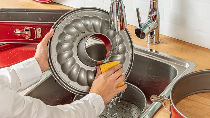Osoba myje formę do ciasta gąbką pod bieżącą wodą w zlewie kuchennym. Obok leżą inne naczynia do pieczenia.