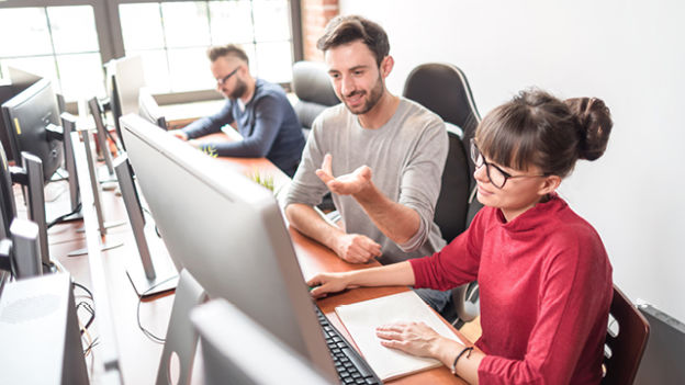 W biurze ludzie pracują przy komputerach. Kobieta w czerwonej bluzce i mężczyzna w szarej koszulce siedzą przy biurku.