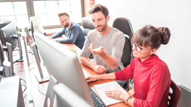 W biurze ludzie pracują przy komputerach. Kobieta w czerwonej bluzce i mężczyzna w szarej koszulce siedzą przy biurku.