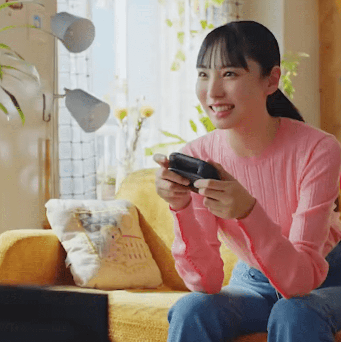 Kobieta w różowym swetrze siedzi na żółtej kanapie i gra na konsoli. W tle okno, lampy i rośliny.