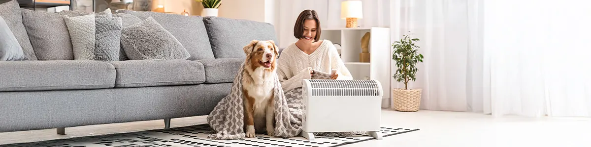 Kobieta z psem siedzą przed małym białym grzejnikiem elektrycznym w jasnym salonie.