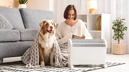 Kobieta z psem siedzą przed małym białym grzejnikiem elektrycznym w jasnym salonie.
