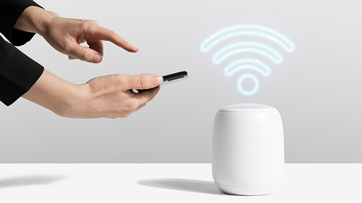 Dłonie trzymają telefon obok białego głośnika z symbolem WiFi. Tło jest jasnoszare, a powierzchnia pod spodem biała.