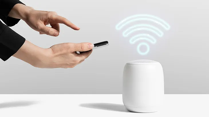 Dłonie trzymają telefon obok białego głośnika z symbolem WiFi. Tło jest jasnoszare, a powierzchnia pod spodem biała.