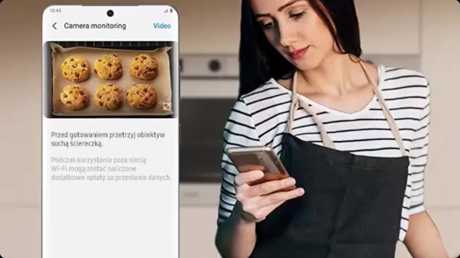 Kobieta w fartuchu sprawdza smartfon w kuchni; na ekranie aplikacja ‘Camera monitoring’ z podglądem z piekarnika – sześć ciastek na blasze i wskazówka o przetarciu obiektywu.
