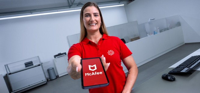 Kobieta w czerwonej koszulce trzyma tablet z logo McAfee w biurze. W tle biurko z klawiaturą i myszą.