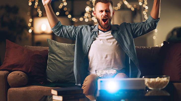 Mężczyzna z uniesionymi rękami siedzi na kanapie. Przed nim projektor, miska z popcornem. W tle dekoracyjne lampki.