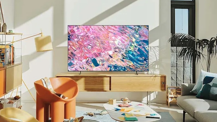 Salon z telewizorem, fotelem i kanapą. Okno z rośliną w tle. Na stole leżą przedmioty. Wnętrze jest jasne.