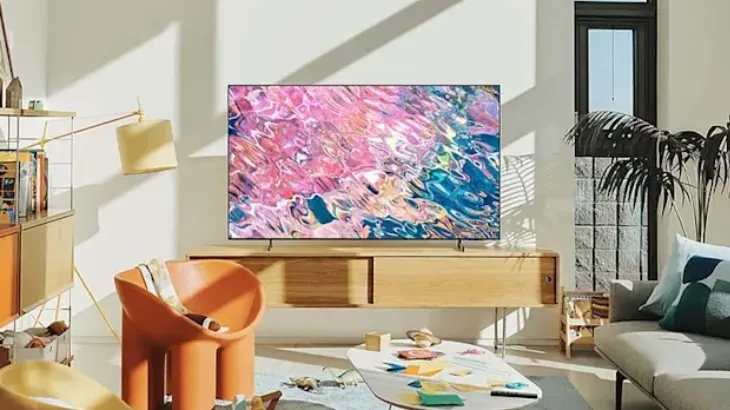 Salon z telewizorem, fotelem i kanapą. Okno z rośliną w tle. Na stole leżą przedmioty. Wnętrze jest jasne.