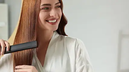 Kobieta w białym szlafroku prostuje włosy prostownicą. W tle lustro, półka i drabina dekoracyjna.