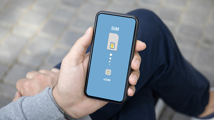 Telefon w dłoni siedzącego człowieka, na ekranie wizualizacje karty SIM i czipa eSIM.