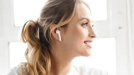 Kobieta z białymi słuchawkami bezprzewodowymi patrzy w prawo, okno w tle.