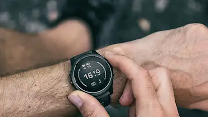 Osoba dotyka zegarka na nadgarstku. Zegarek pokazuje godzinę 16:19. Tło jest ciemne i nieostre.