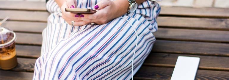 Osoba w pasiastym stroju siedzi na drewnianej ławce, trzymając telefon. Obok stoi napój i leży biały przedmiot.