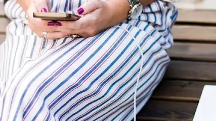 Osoba w pasiastym stroju siedzi na drewnianej ławce, trzymając telefon. Obok stoi napój i leży biały przedmiot.