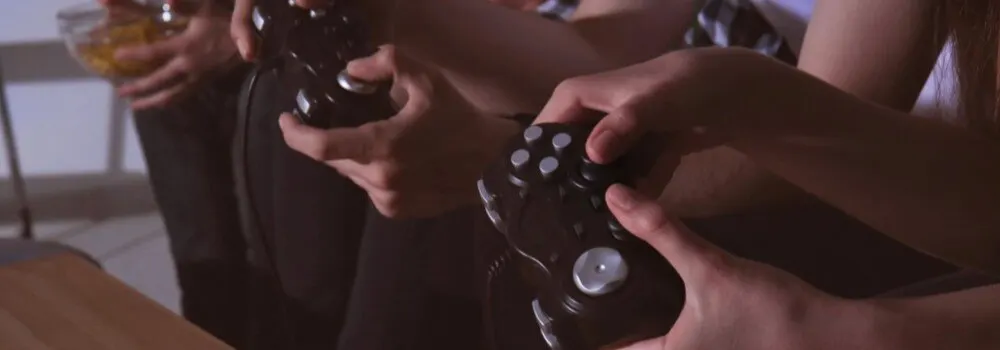 Bliski plan na ręce trzymające czarne kontrolery do gier, siedzące osoby, jedna z miską przekąsek.