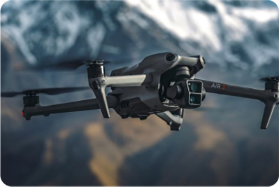 Czarny dron z kamerą w locie, na tle gór z białym śniegiem i brązowymi zboczami.