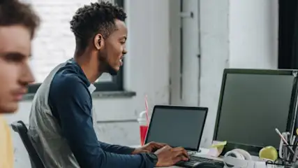 Mężczyźni pracują przy biurkach z laptopami i komputerami. Na biurkach znajdują się napoje, jabłka i akcesoria biurowe.