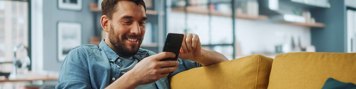 Uśmiechnięty mężczyzna w koszuli siedzi na żółtej kanapie, używając smartfona w salonie.