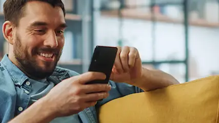 Uśmiechnięty mężczyzna w koszuli siedzi na żółtej kanapie, używając smartfona w salonie.