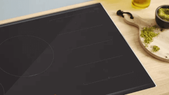 Czarny blat kuchenny obok deski do krojenia z ziołami i miseczki, na drewnianym blacie. W tle butelka z płynem.