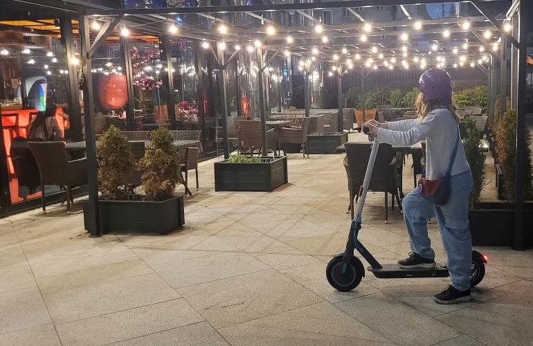 Osoba na hulajnodze w tle oświetlonej kawiarni ze stolikami i krzesłami. Nad głową wiszą światełka.