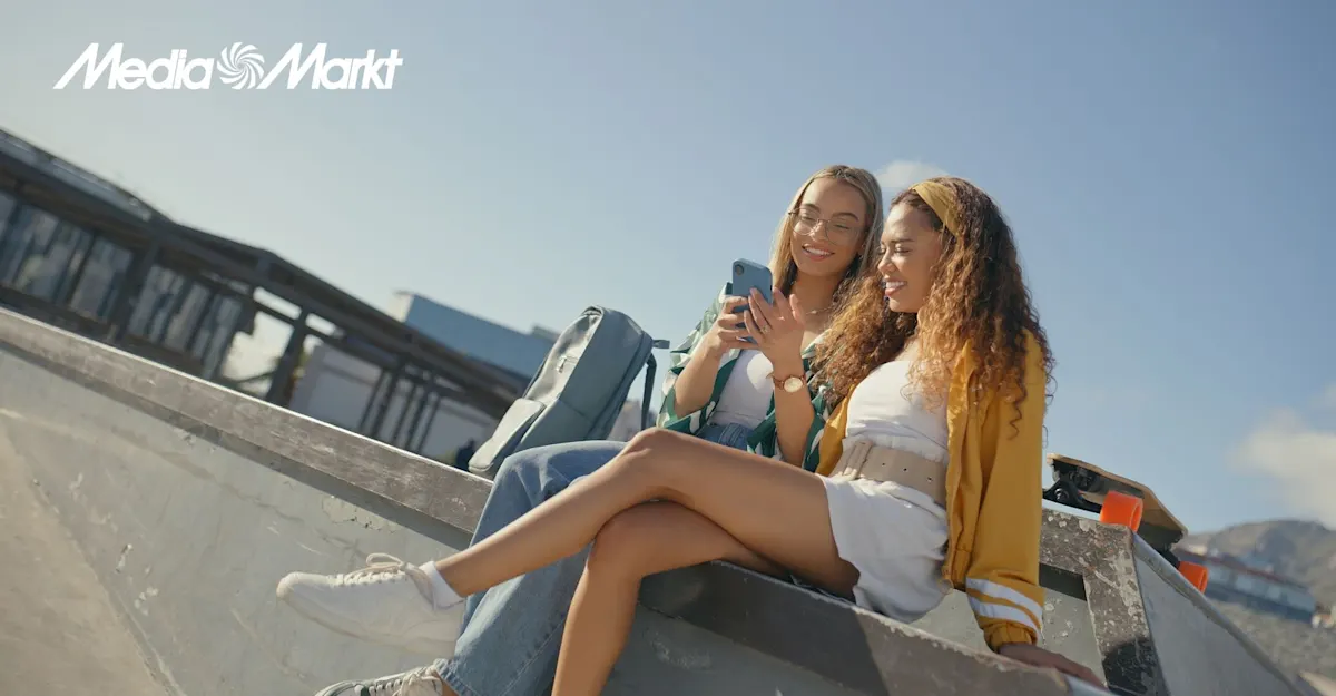 Dwie kobiety siedzą na rampie, patrzą na telefon. Obok leży deskorolka, w tle widać budynki i błękitne niebo.