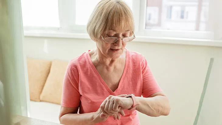 Kobieta w okularach patrzy na zegarek na ręce, ubrana w różową koszulkę. W tle okno i beżowe poduszki na kanapie.