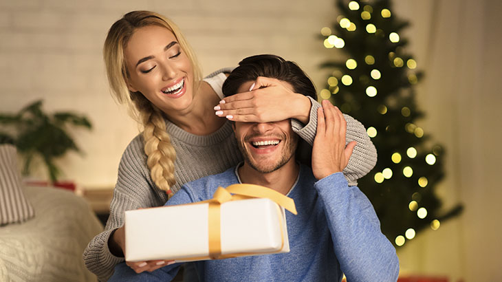 Kobieta zasłania oczy mężczyźnie i wręcza mu prezent. W tle choinka.