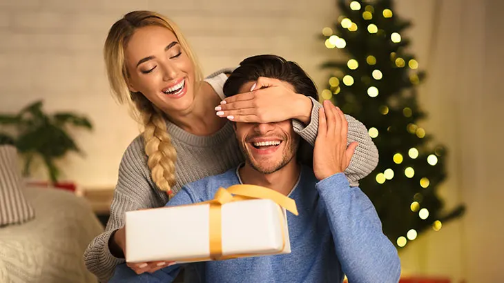 Kobieta zasłania oczy mężczyźnie i wręcza mu prezent. W tle choinka.